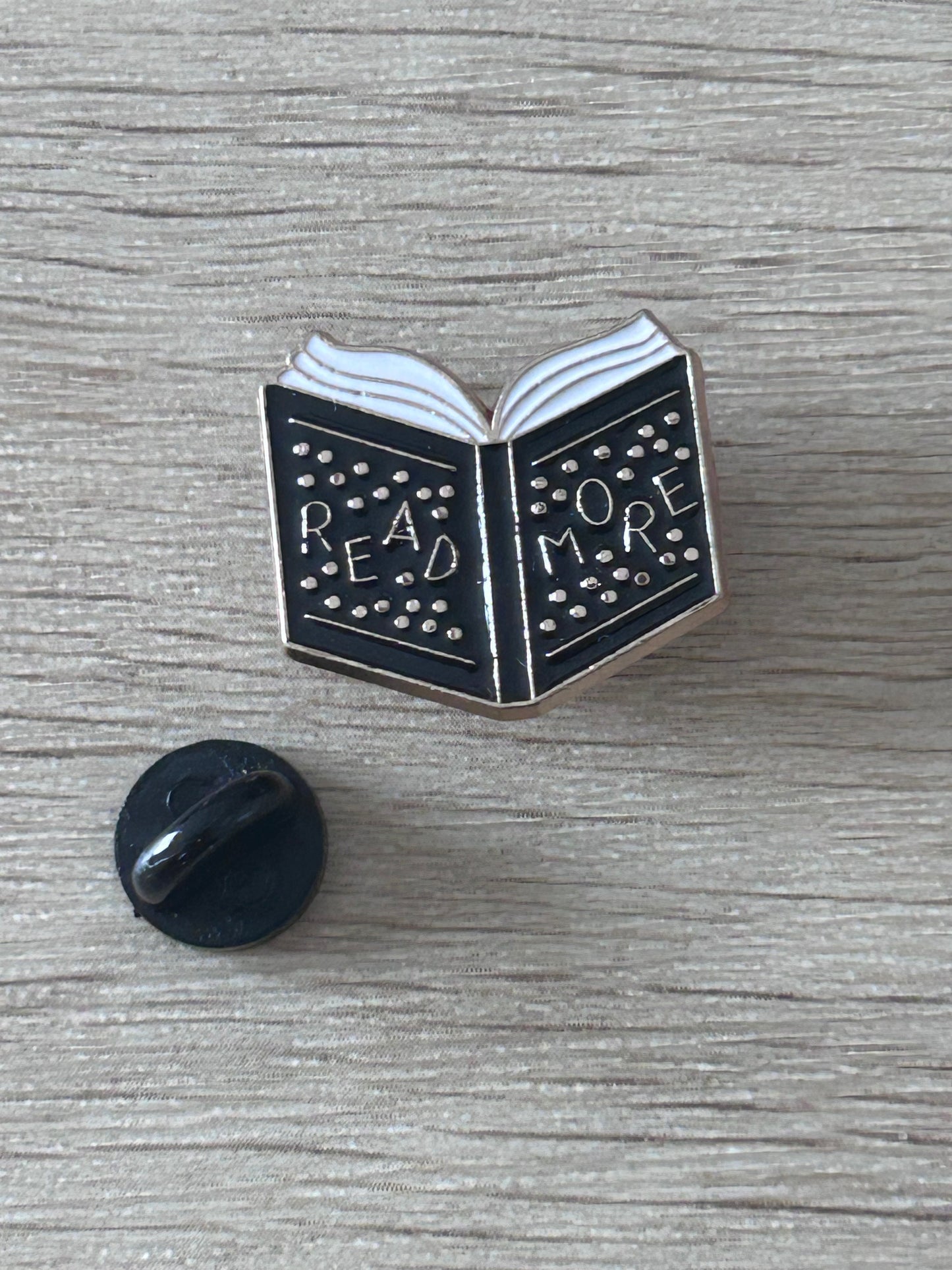 Goupille en forme de livre ouvert noir et blanc, texte perforé "READ MORE".