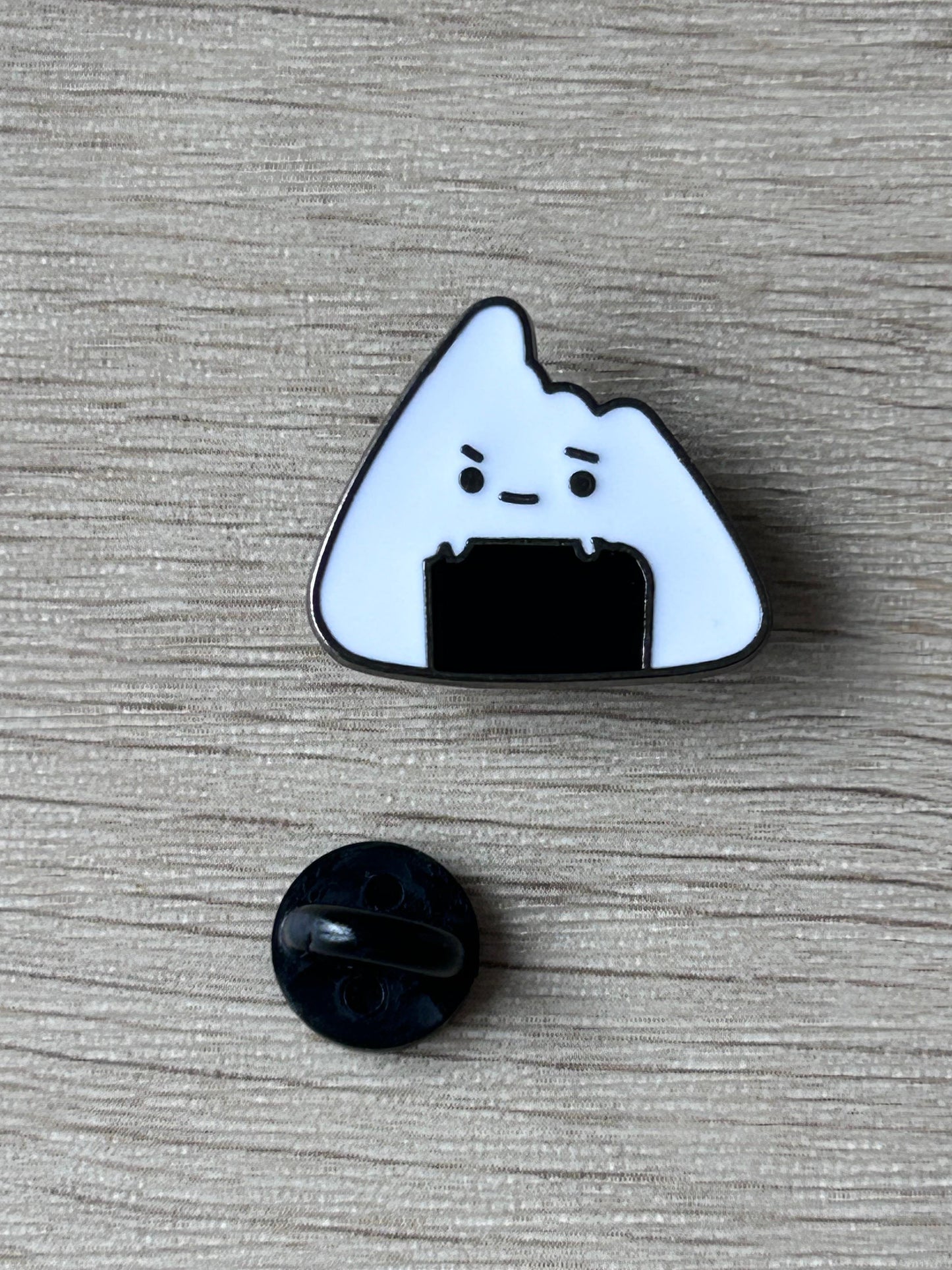 Épingle onigiri blanche au visage grognon, contour foncé; disque noir séparé.