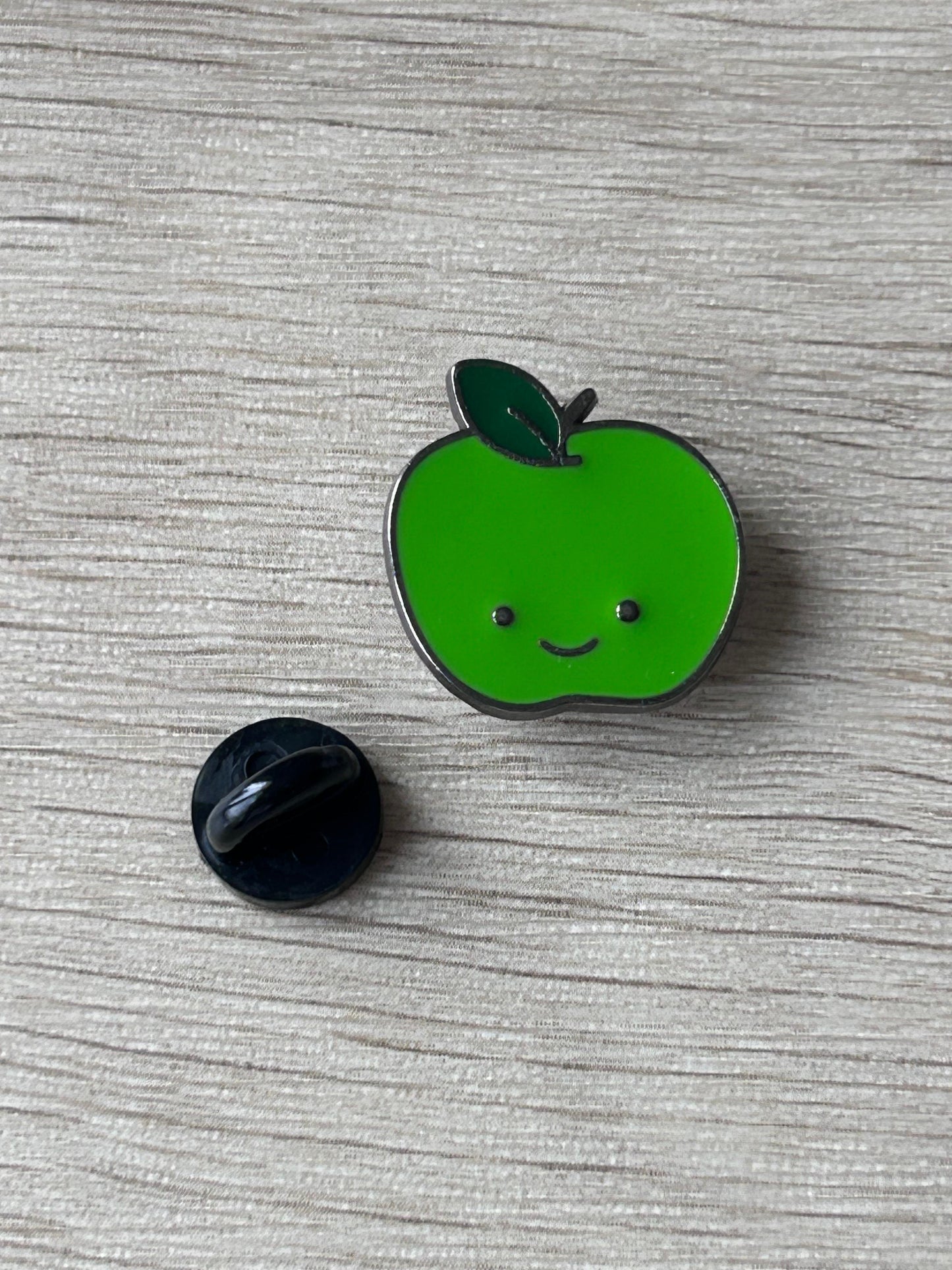 Épingle pomme verte souriante, dos avec tige noire et bord argenté, feuille sombre.
