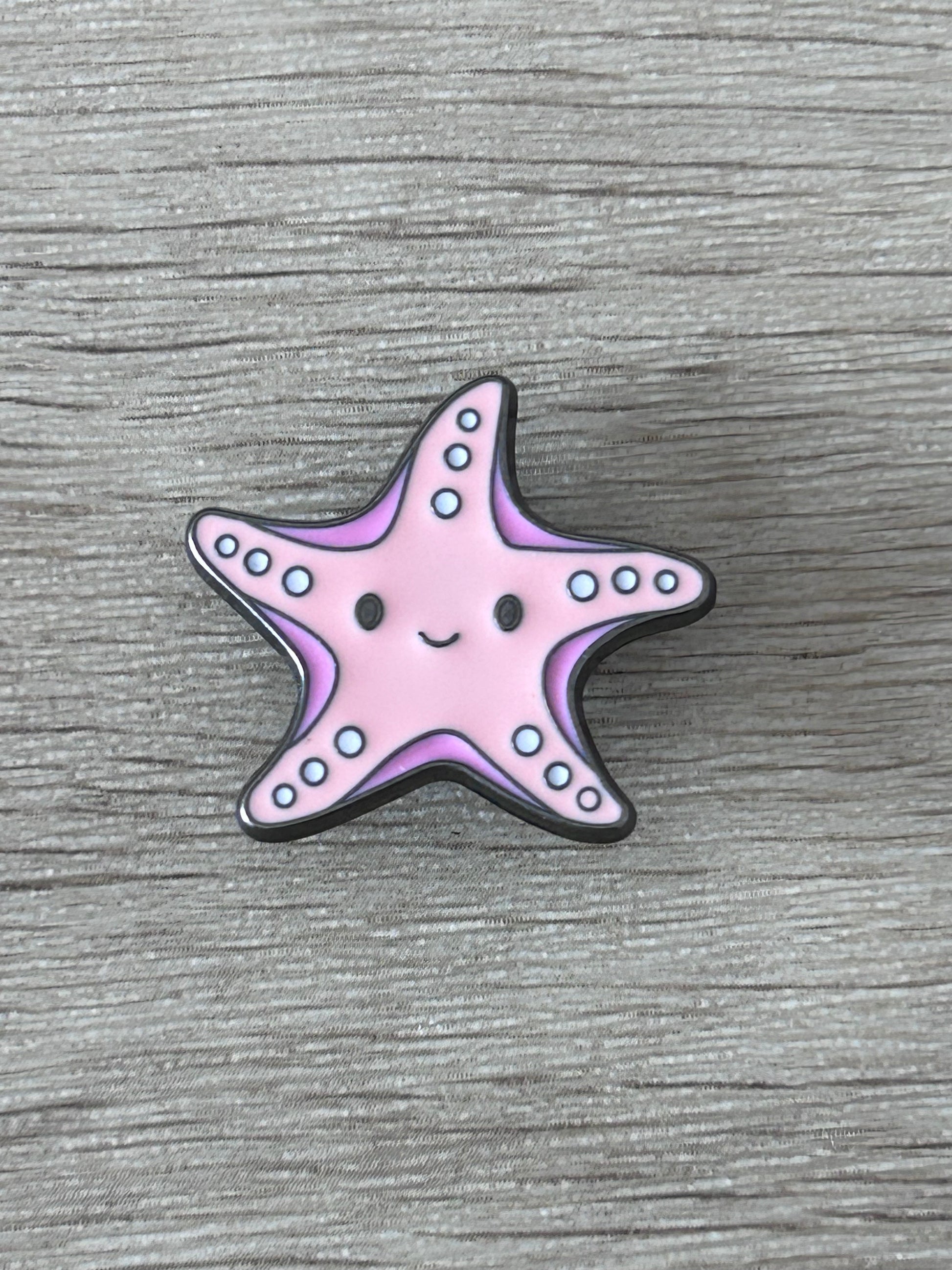 Pin's en forme d’étoile de mer kawaii rose, sourire et points bleus, contour noir.