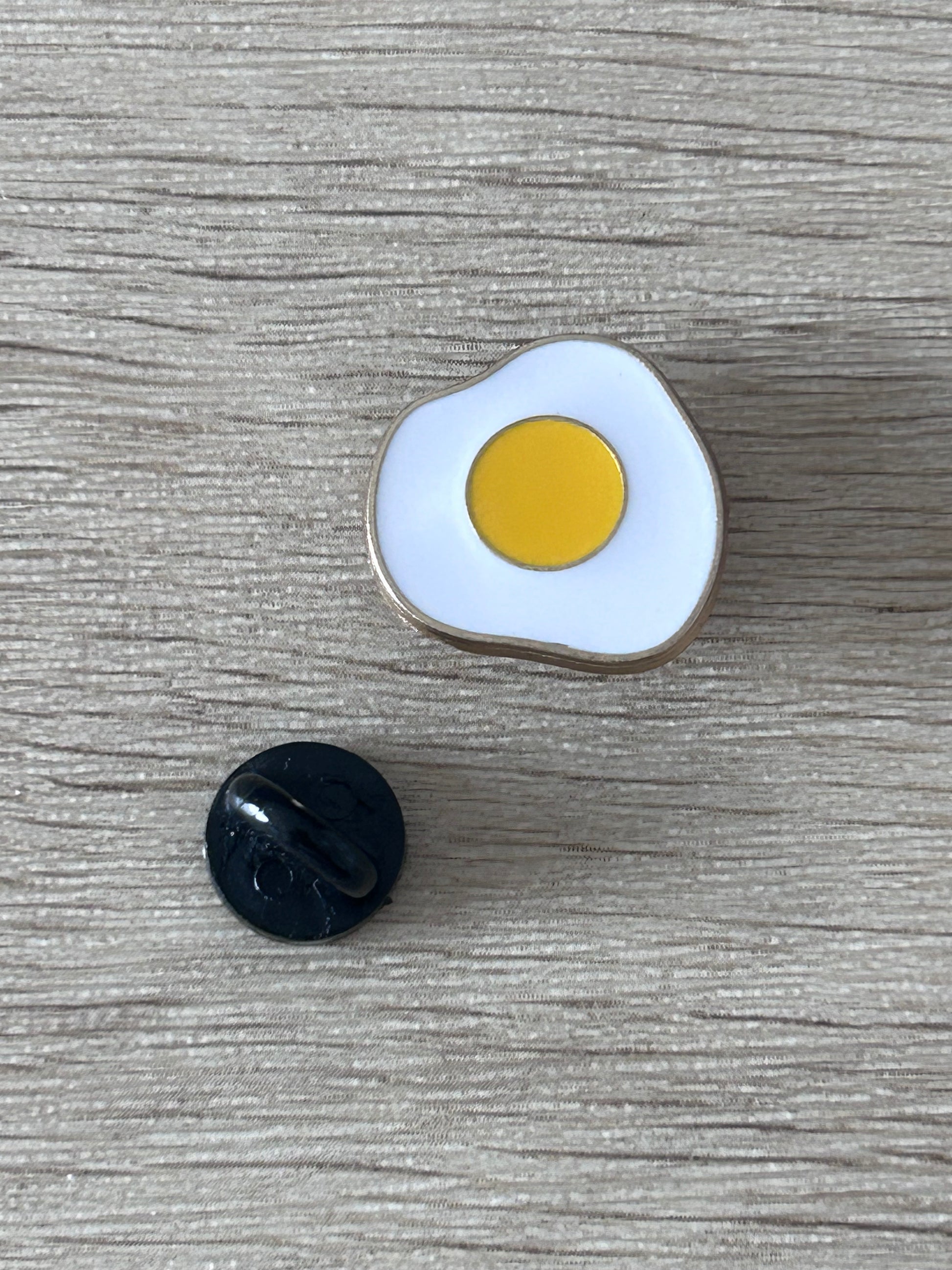 Épingle émaillée en forme d’œuf au plat, contour argent, jaune sur fond blanc.