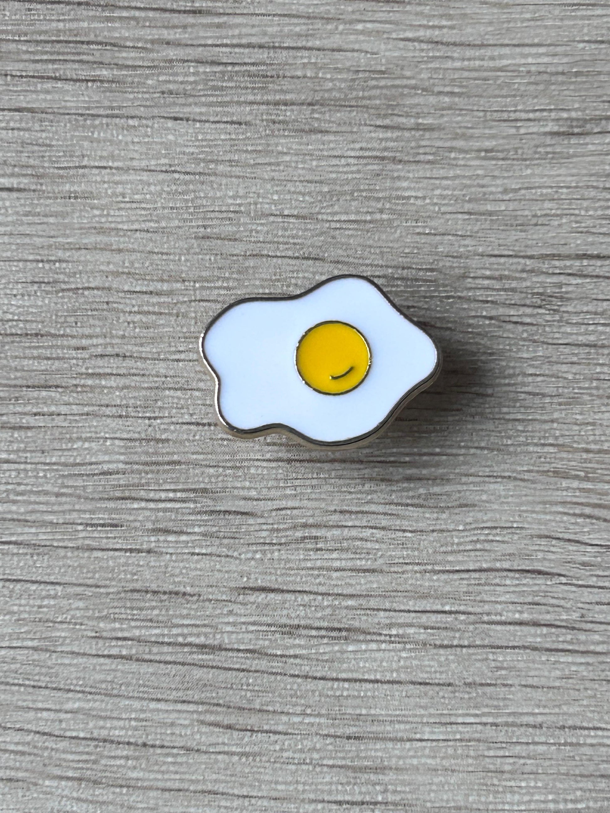 Broche en forme d’œuf au plat émaillé, blanc et jaune, contours bleutés.