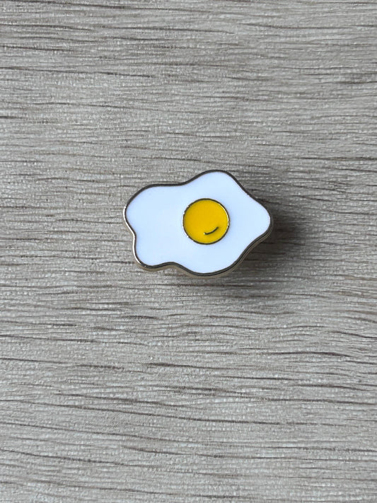 Broche en forme d’œuf au plat émaillé, blanc et jaune, contours bleutés.