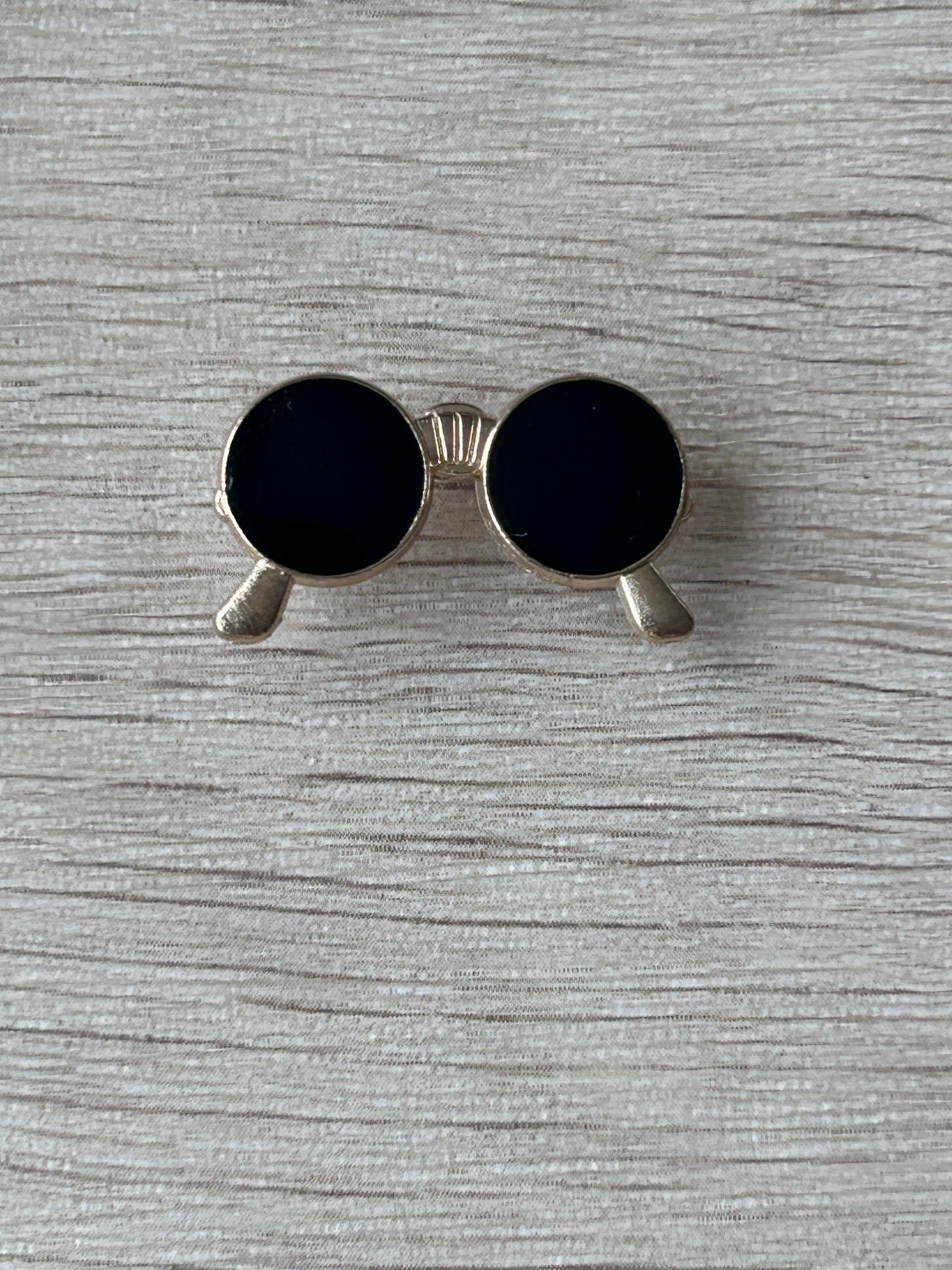 Broche lunettes rondes noires : monture argentée et embouts gris, verres noirs.