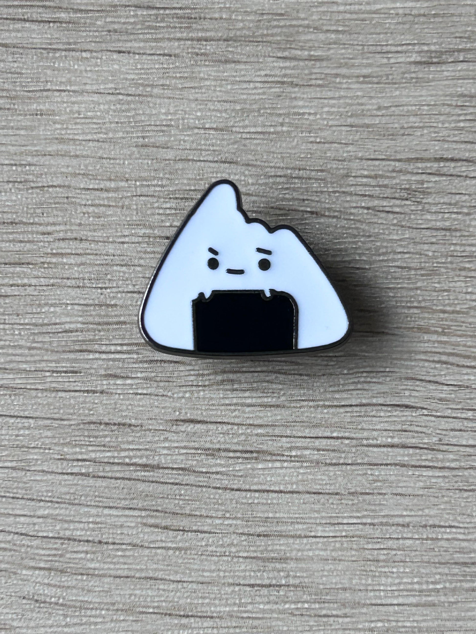 Épingle en forme d’onigiri blanc, visage grognon et contour bleu foncé.