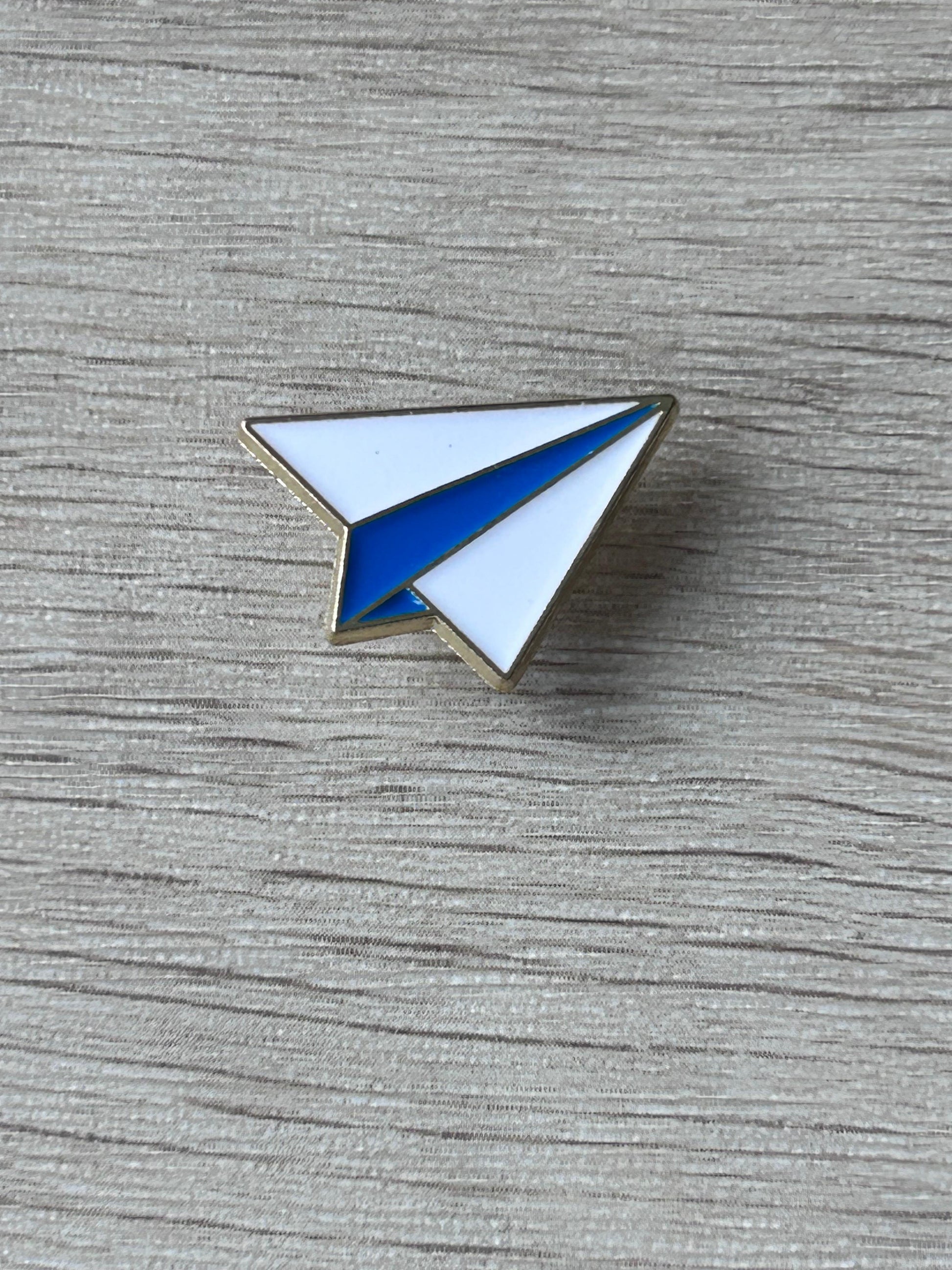 Pin avion en papier, triangles blanc et bleu bordés d’or.