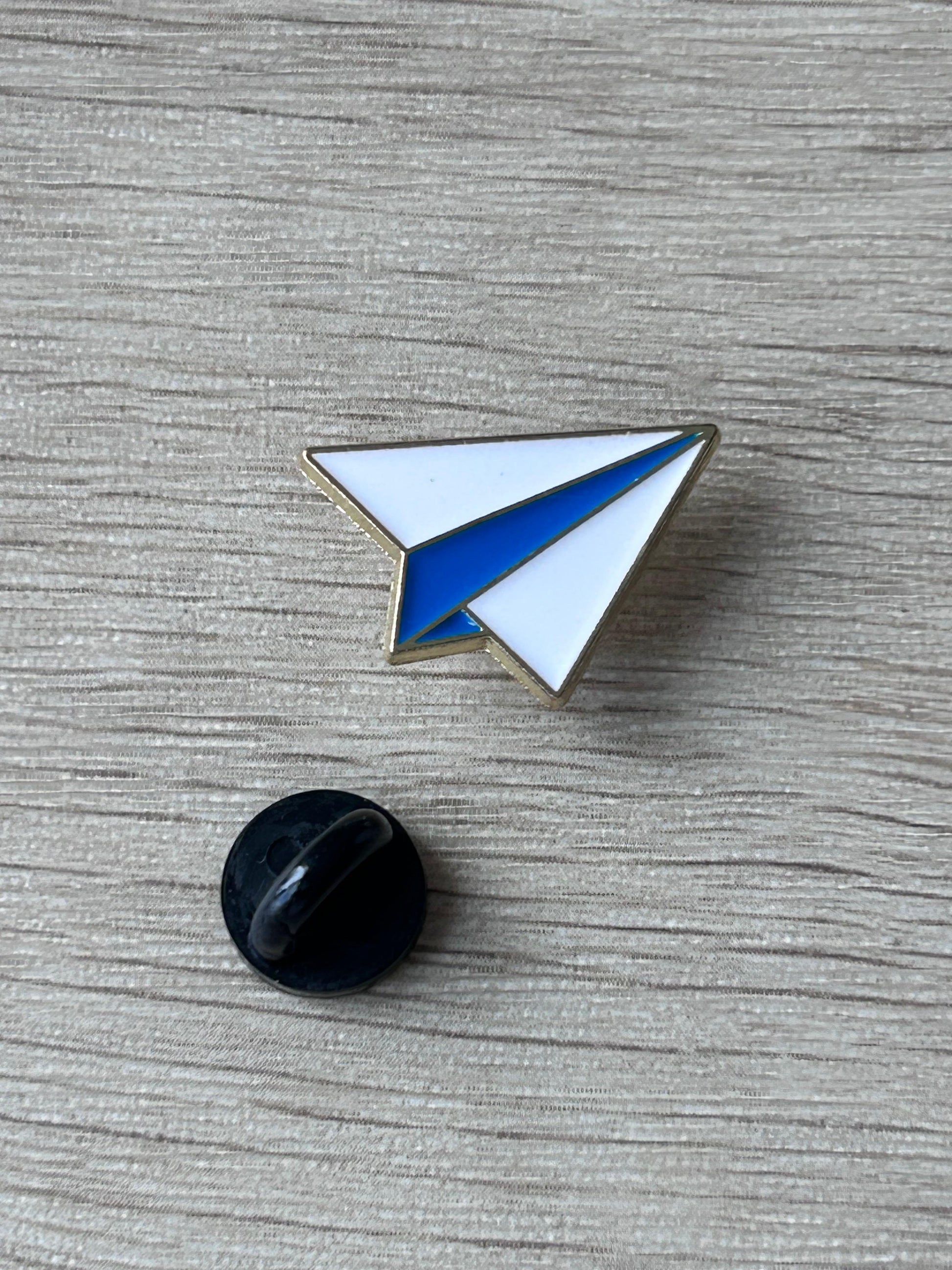 Pin avion en papier avec attache noire au dos, triangles blanc et bleu bordés d’or.