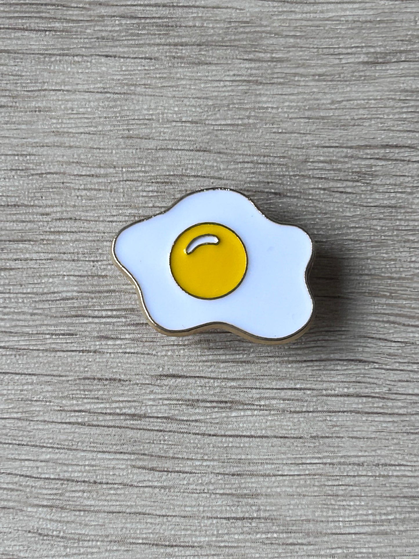 Pin œuf au plat émaillé jaune et blanc à bordure noire.