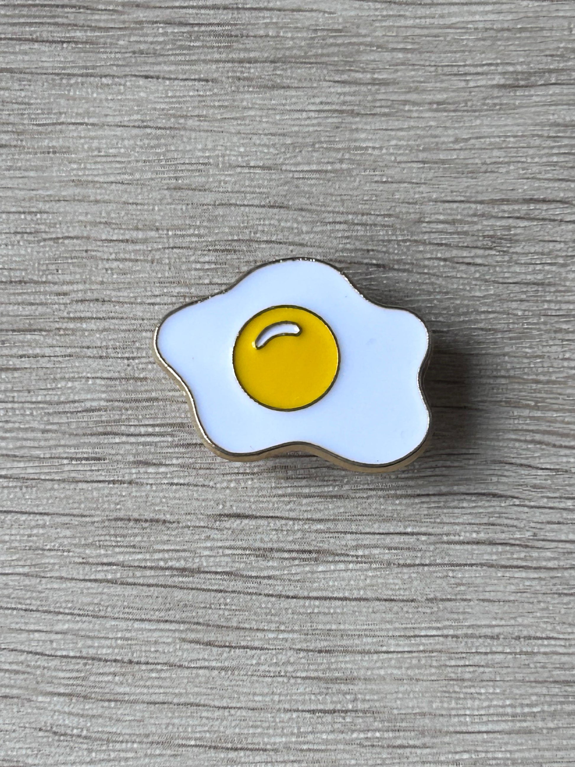 Pin œuf au plat émaillé jaune et blanc à bordure noire.