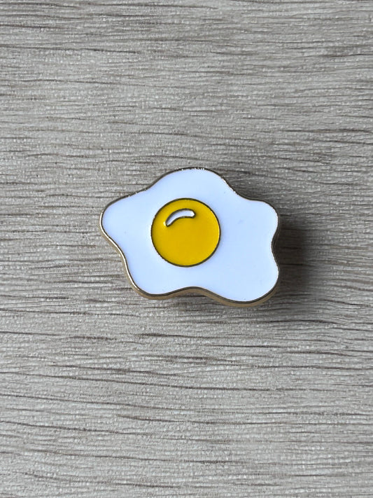 Pin œuf au plat émaillé jaune et blanc à bordure noire.