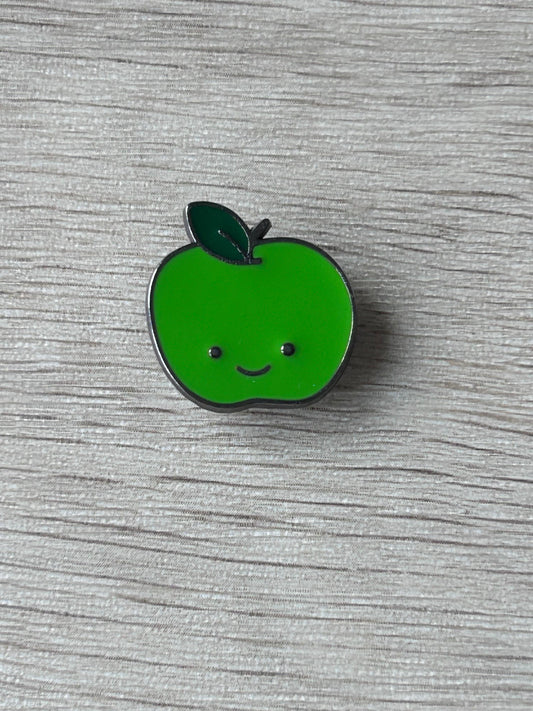 Épingle en forme de pomme verte souriante, bord argenté et feuille sombre.