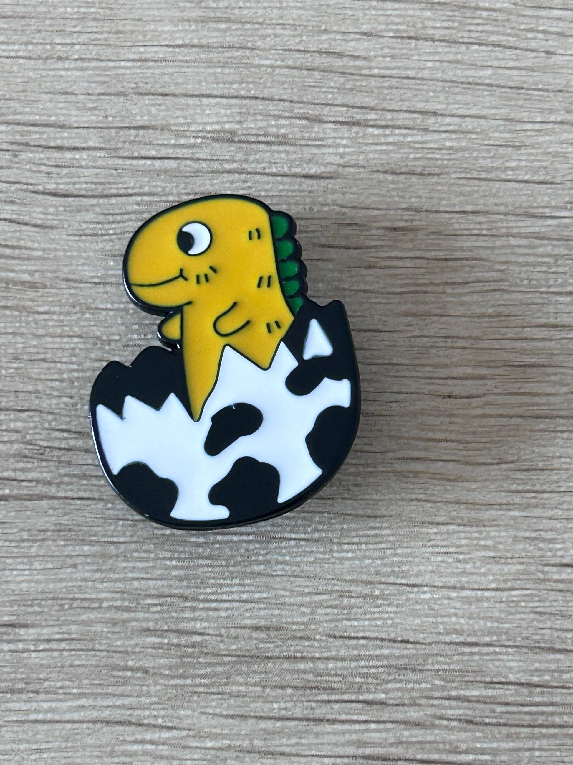 Pin bébé dinosaure jaune-vert, noir et blanc, en forme d’œuf avec visage souriant.