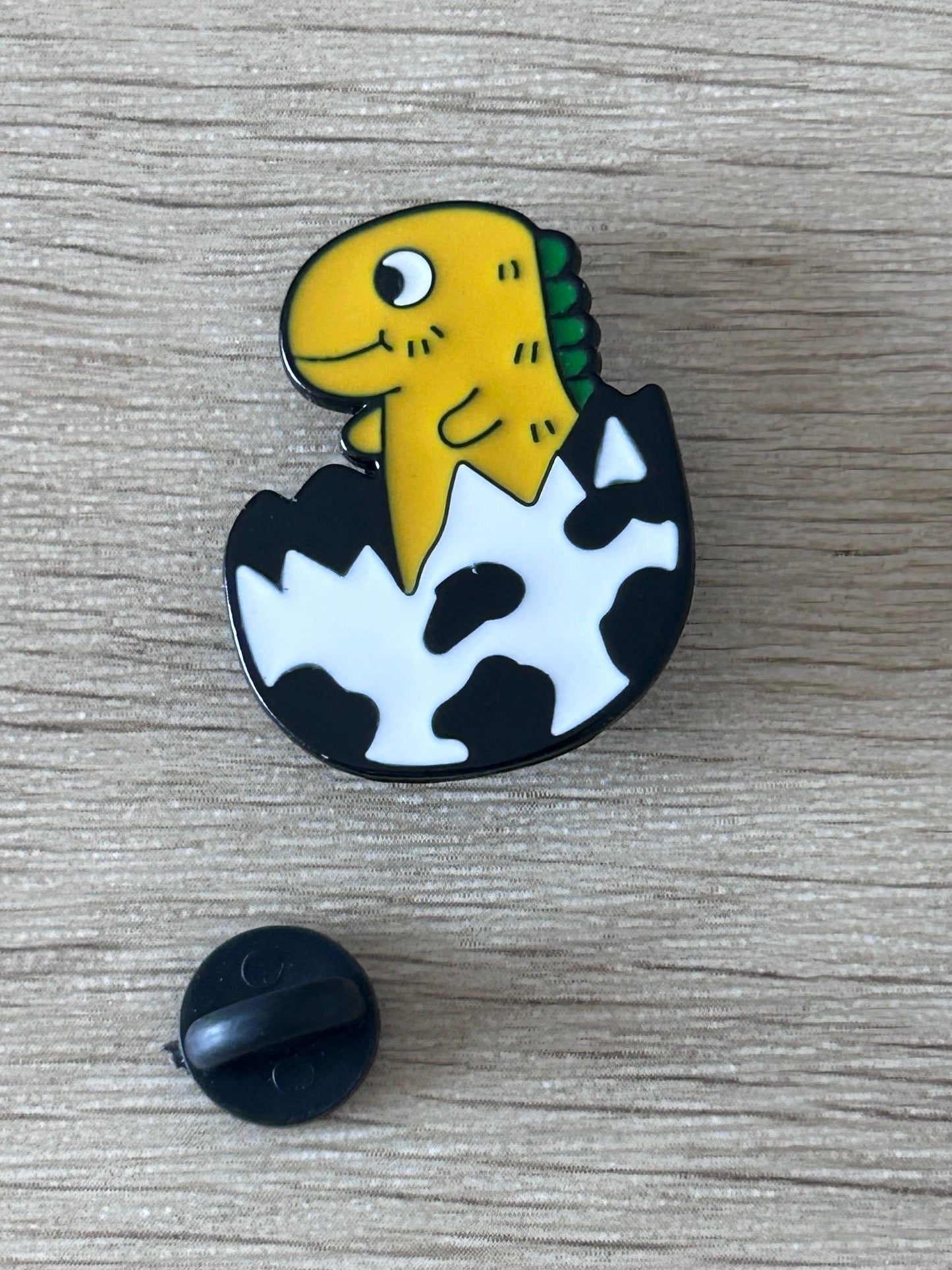 Pin bébé dinosaure jaune-vert noir et blanc, en forme d’œuf, avec support rond noir.