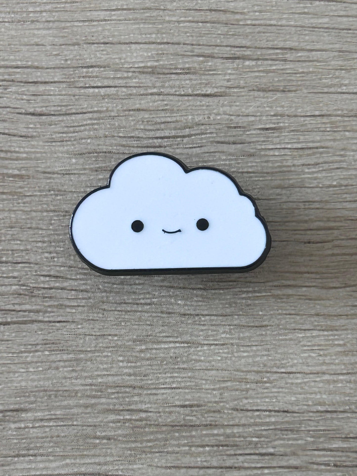 Pin's nuage souriant bleu clair à contour noir, yeux ronds et sourire discret.