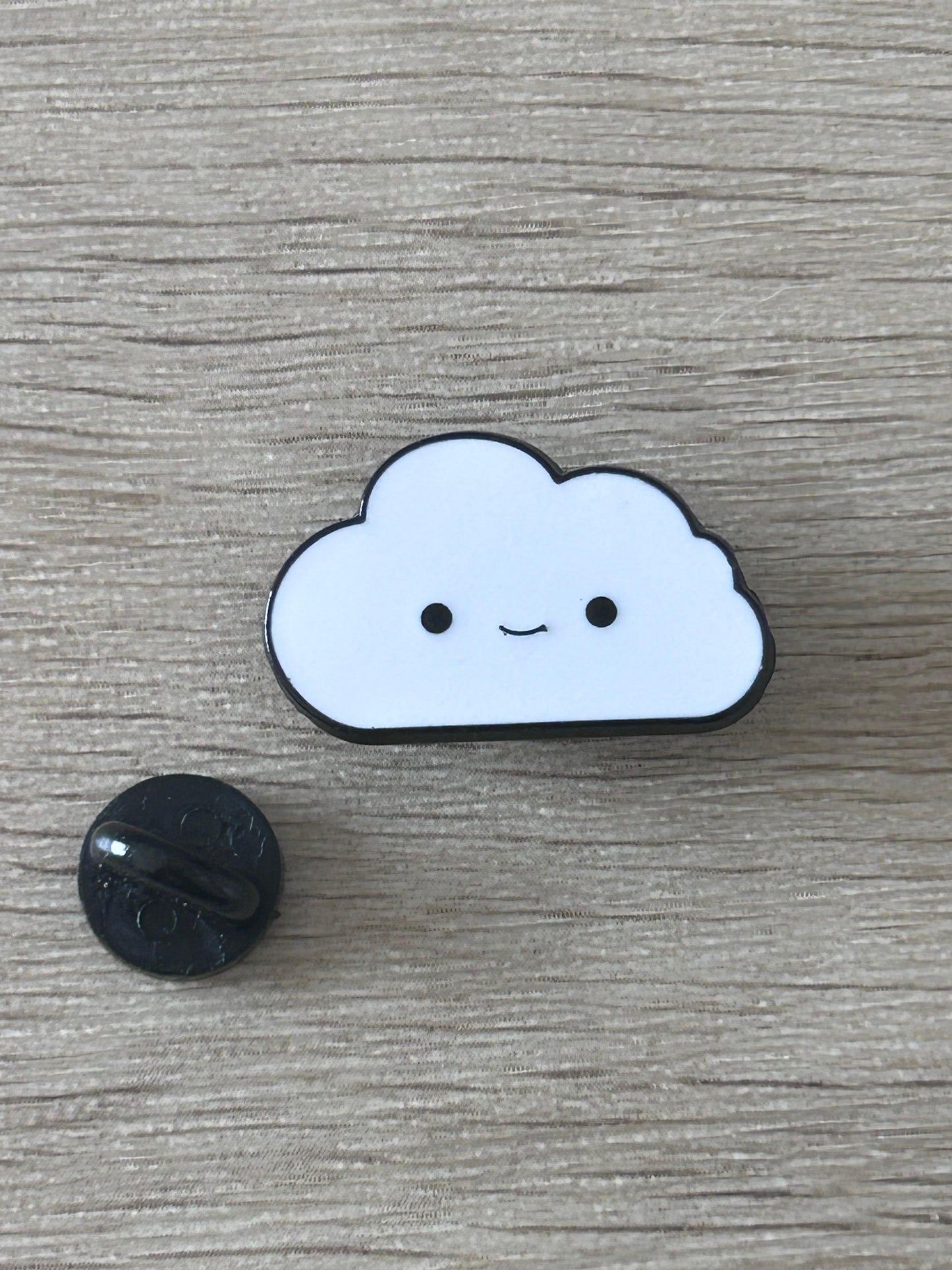 Pin's nuage souriant bleu clair à contour noir, avec attache noire visible.