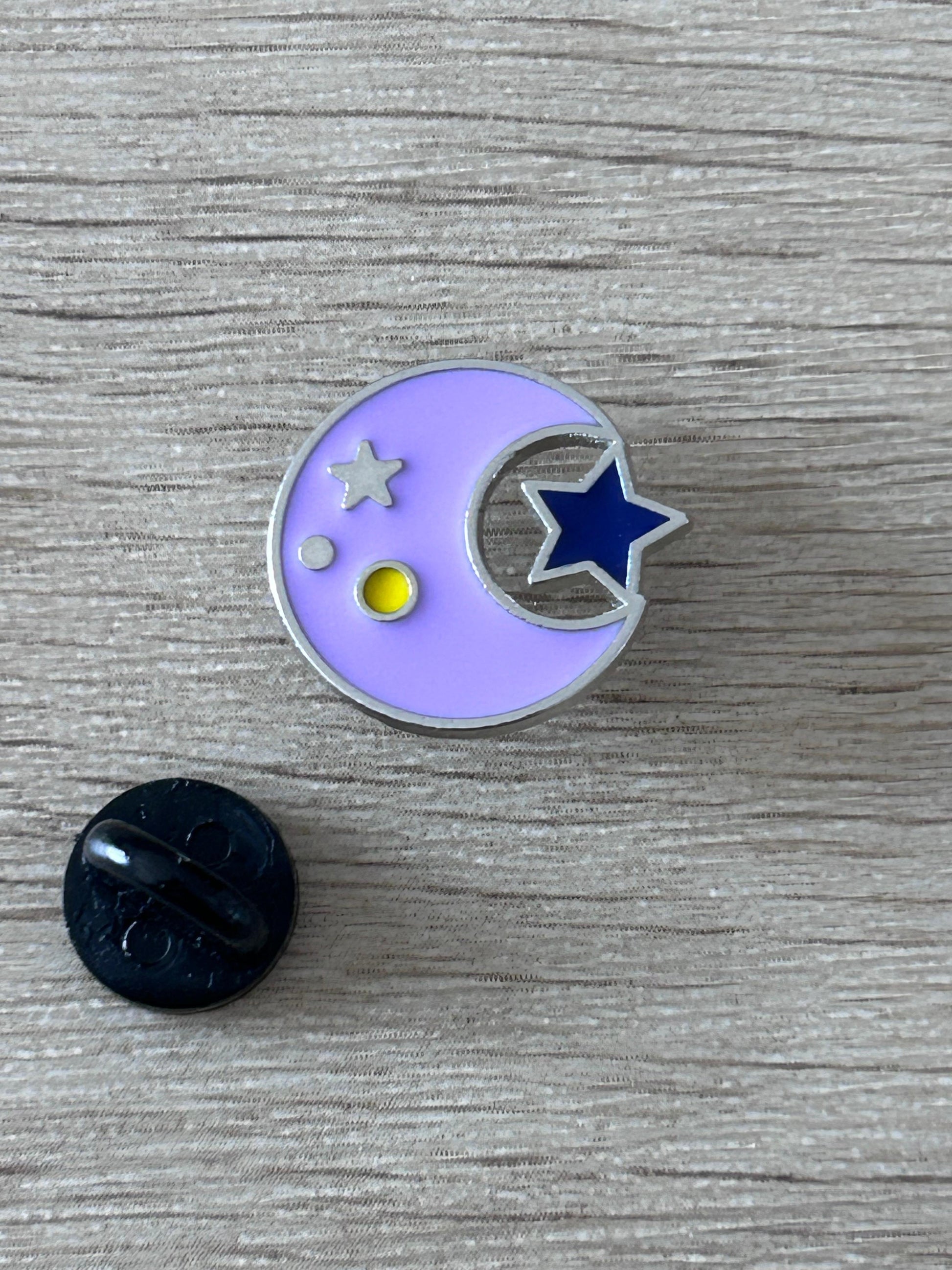 Pin's rond lilas avec lune et étoiles, face avant et attache noire visible.