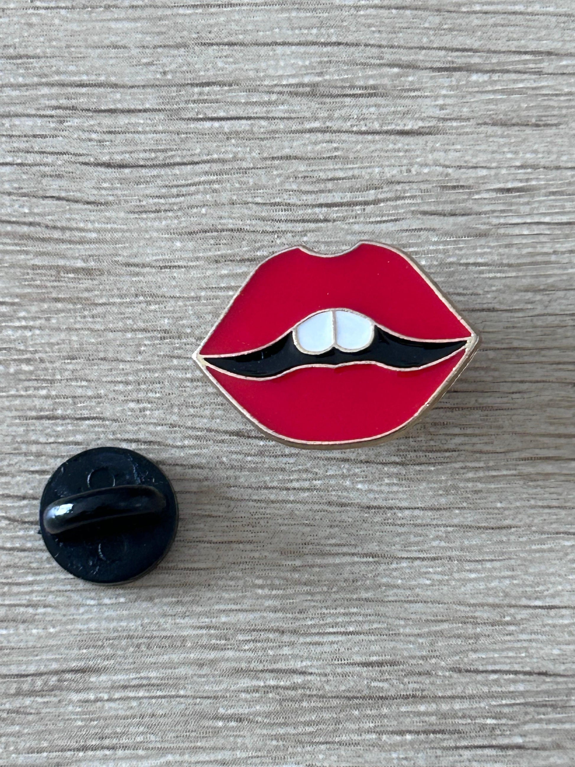 Épingle en forme de lèvres rouges, dents blanches visibles et contour noir.