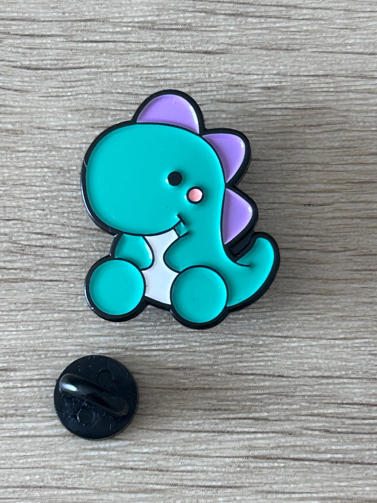 Broche dinosaure bébé kawaii turquoise et violet, contour noir; dos rond noir visible.