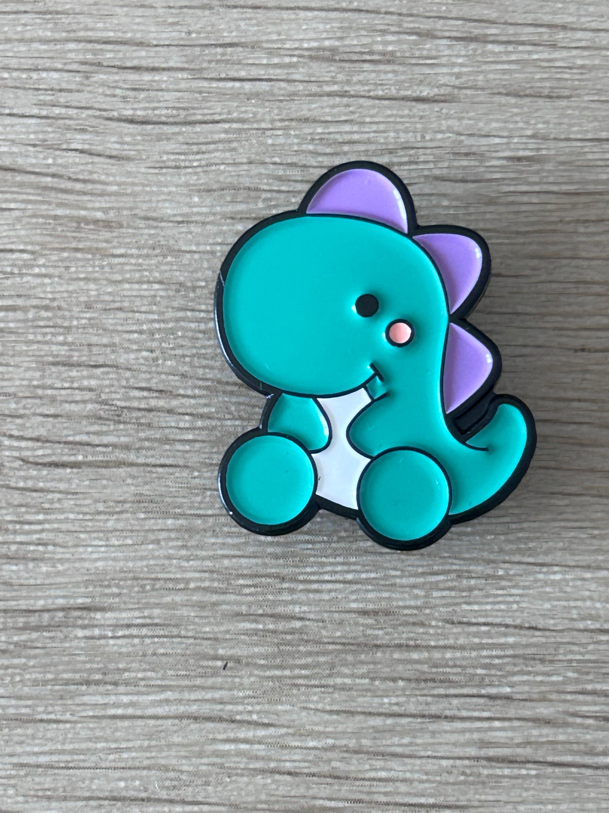 Broche dinosaure bébé kawaii turquoise et violet, contour noir, tête souriante.