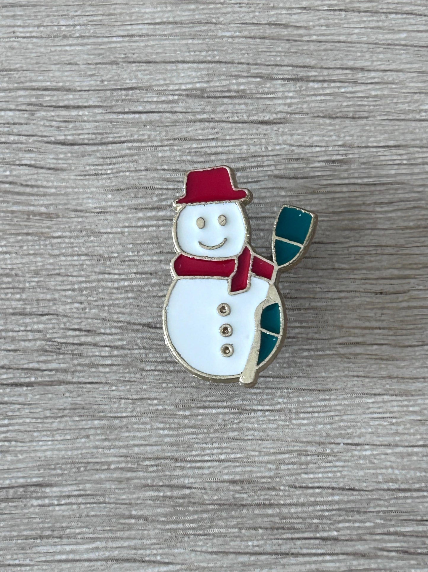 Broche bonhomme de neige émaillée : chapeau rouge, écharpe verte, bonhomme blanc souriant.