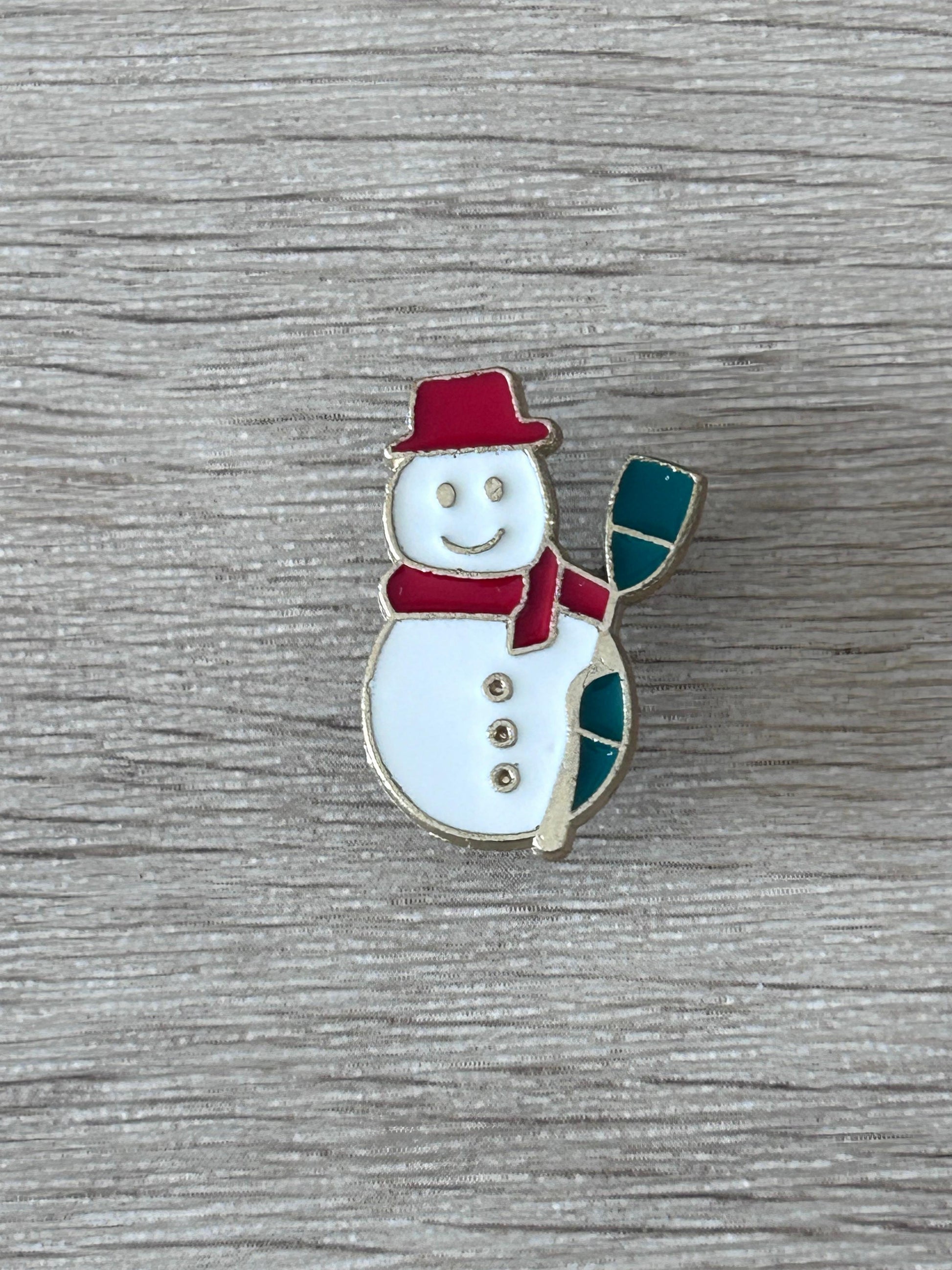 Broche bonhomme de neige émaillée : chapeau rouge, écharpe verte, bonhomme blanc souriant.
