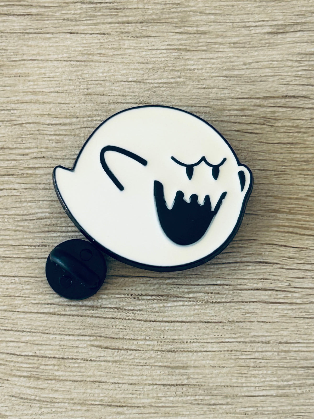 Pin émaillé fantôme blanc, visage noir souriant, attache arrière noire visible.