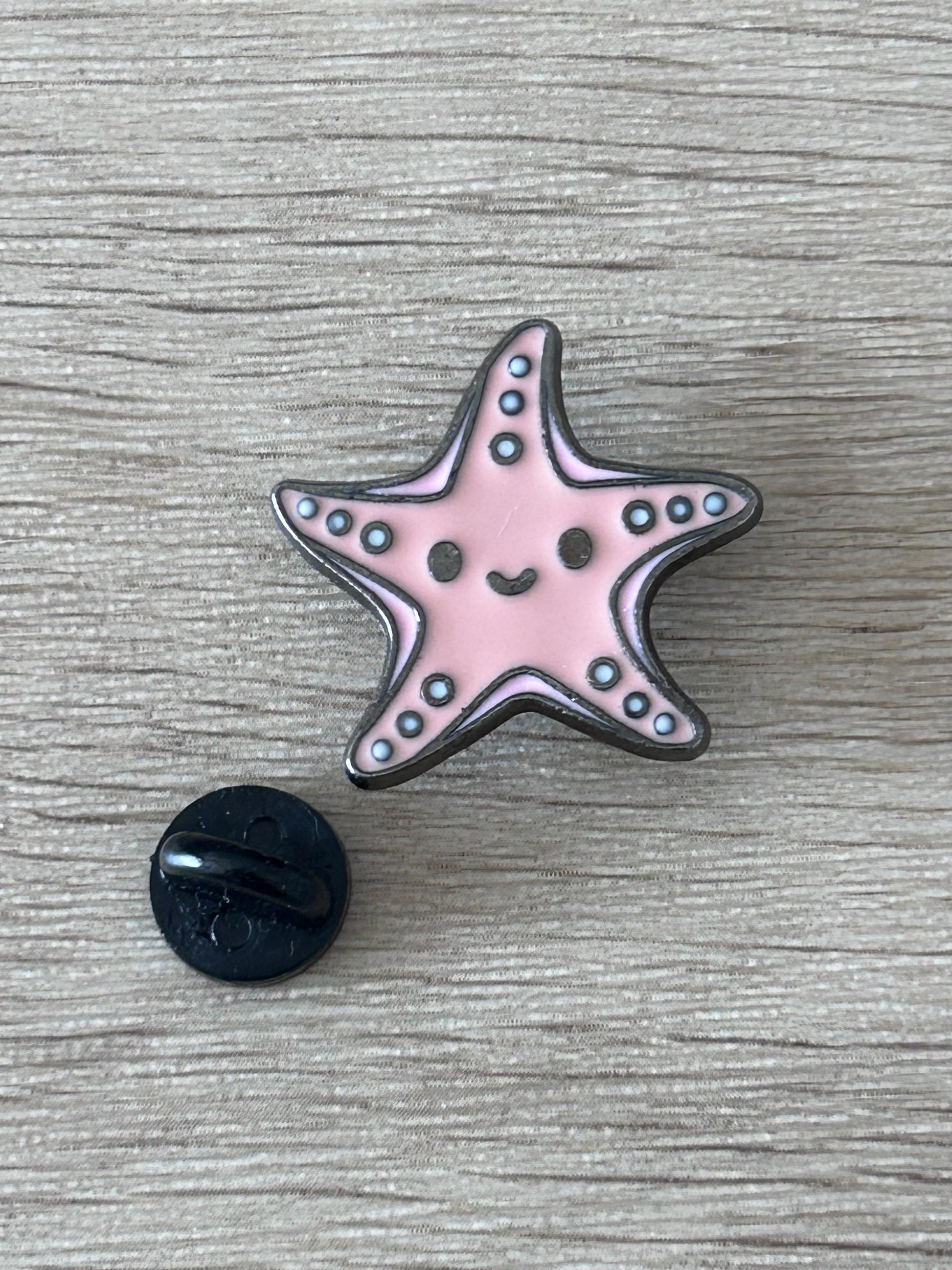 Pin's en forme d’étoile de mer kawaii rose avec dos noir rond, sourire et points bleus.