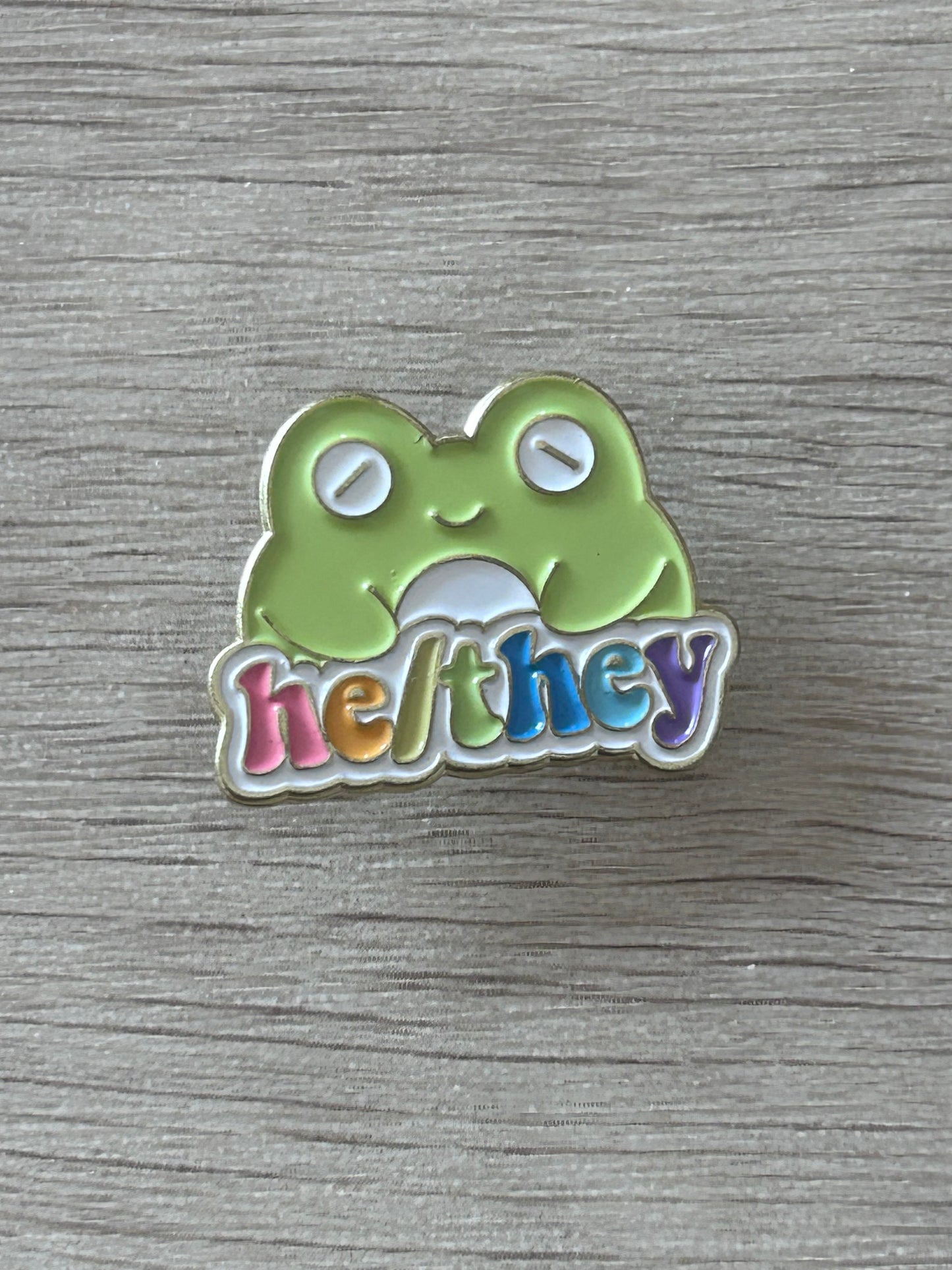 Pin's he/they coloré avec papillon vert sur fond en bois