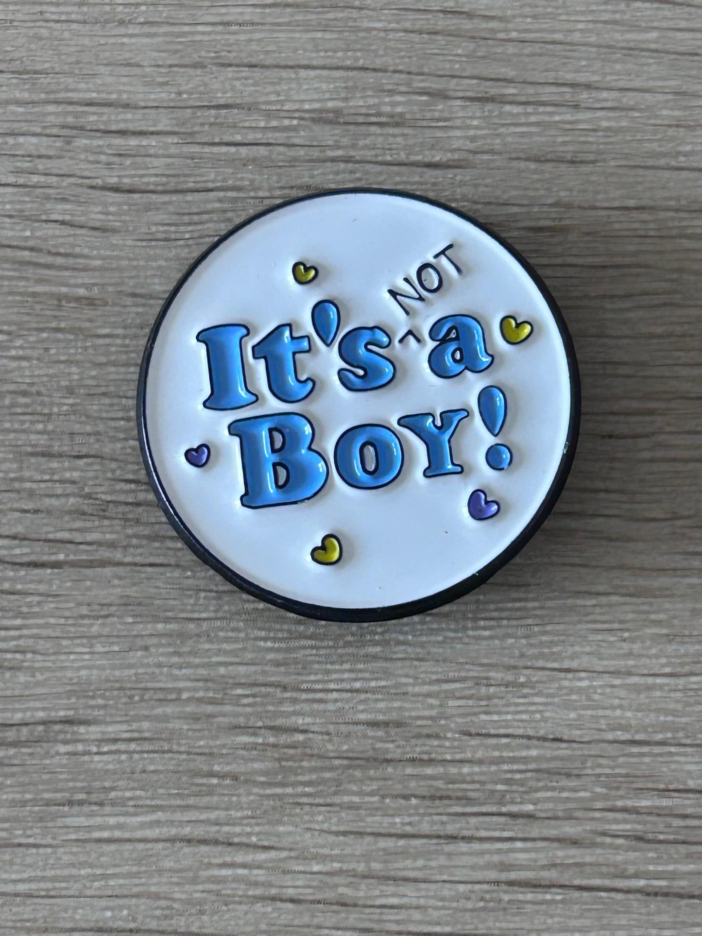 Pin's rond avec inscription « It's NOT a Boy! » bleu sur fond clair, bord noir.
