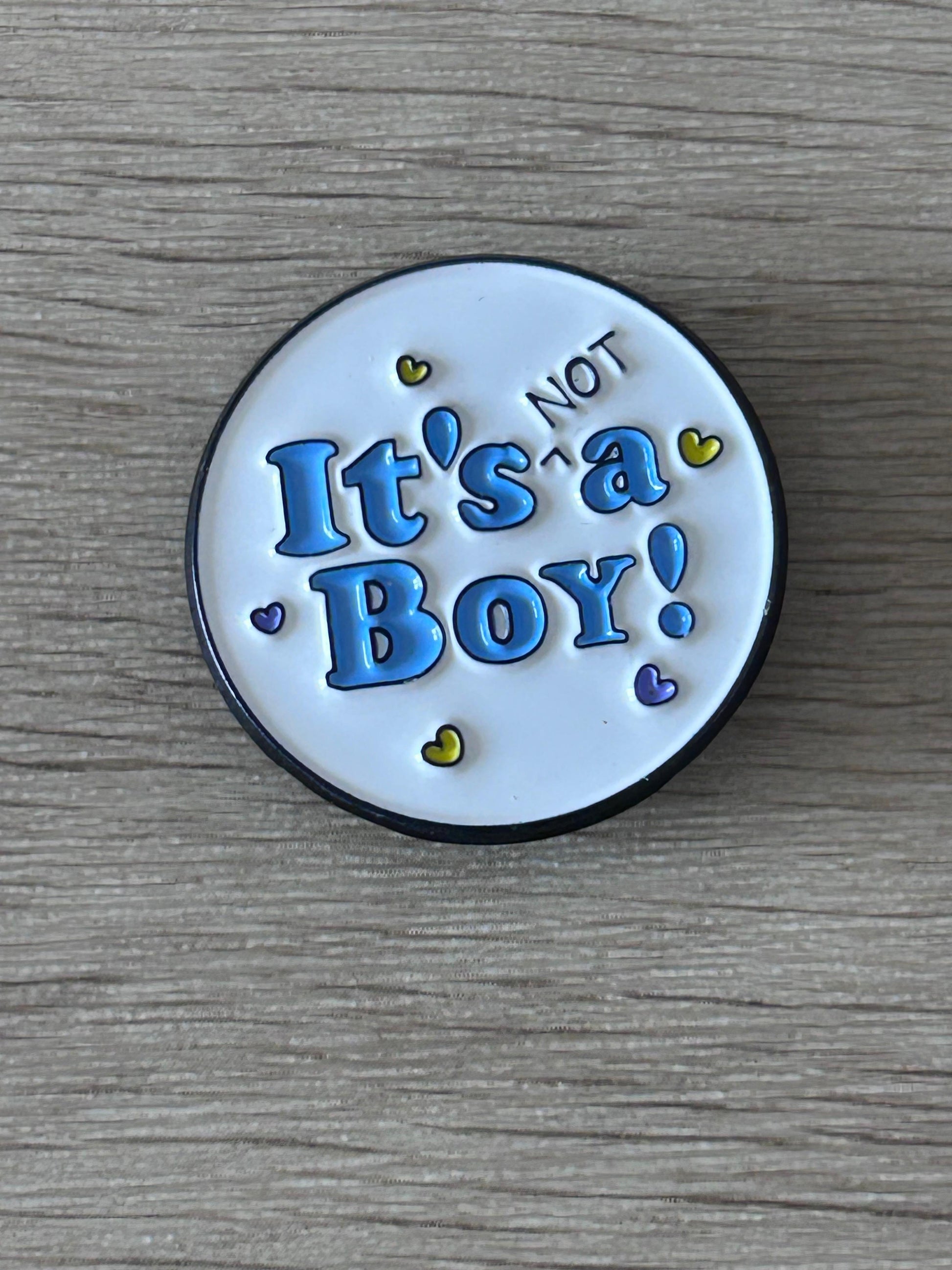 Pin's rond avec inscription « It's NOT a Boy! » bleu sur fond clair, bord noir.