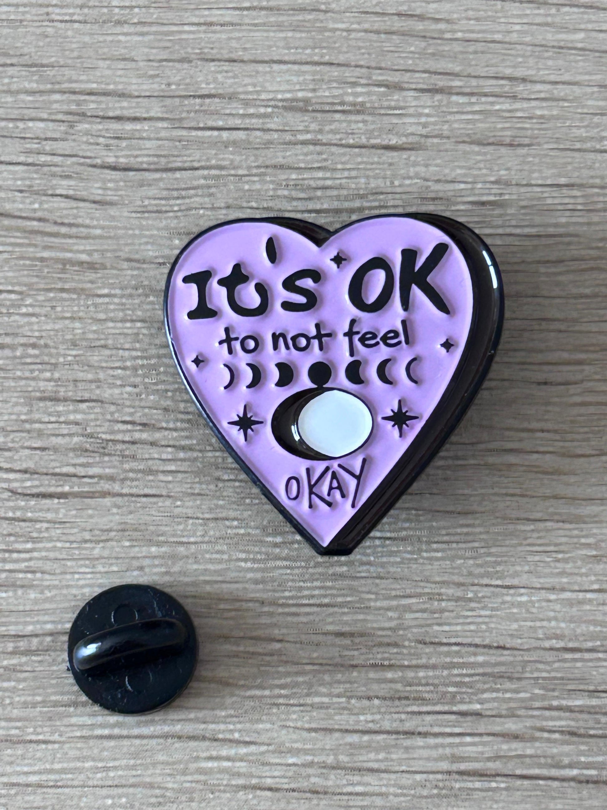 Pin cœur violet « It’s OK »; texte et dos noir avec attache visible.