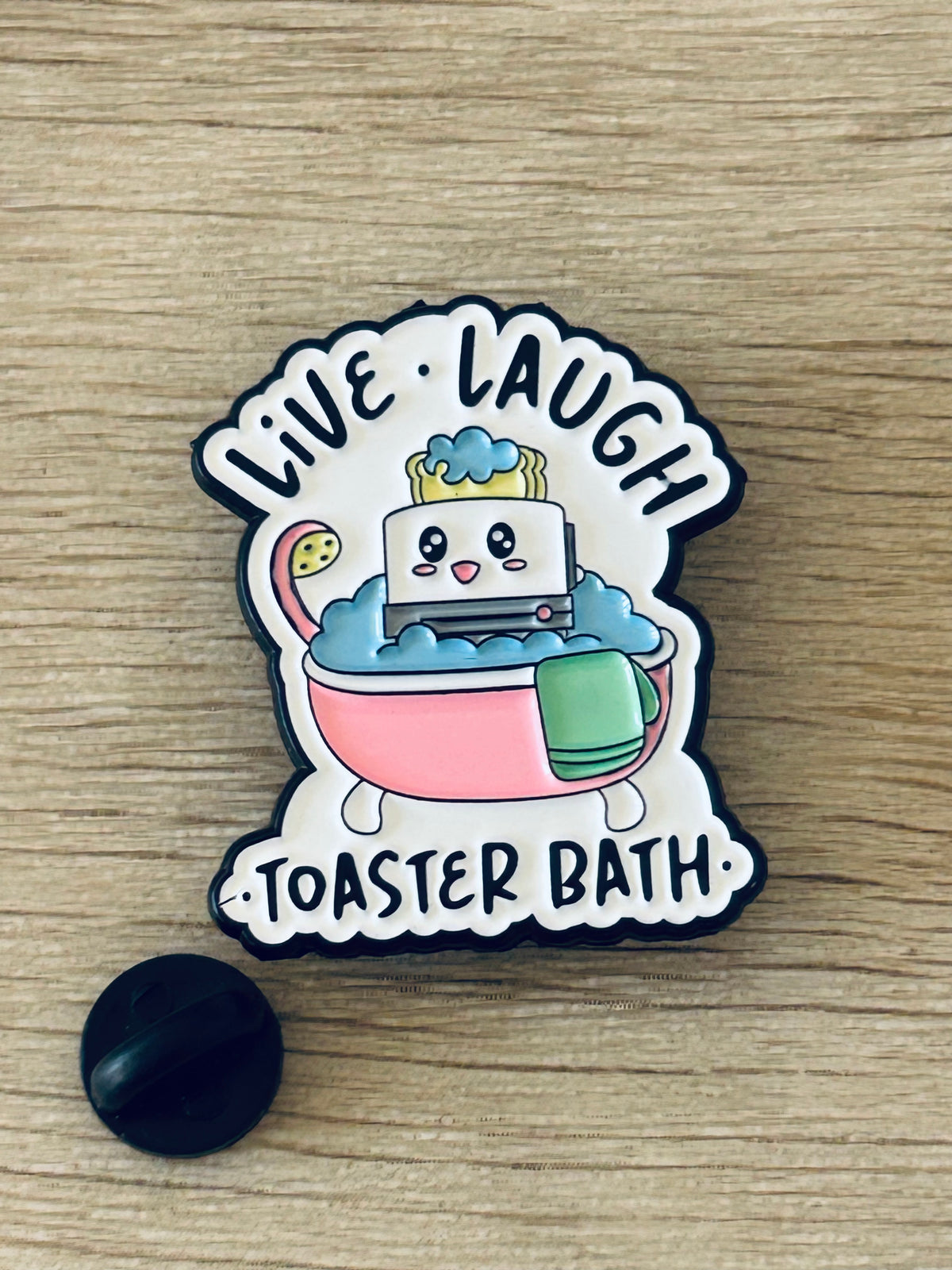Pin émaillé toaster en baignoire, couleur rose et vert, texte “Live Laugh” et “Toastier Bath”.
